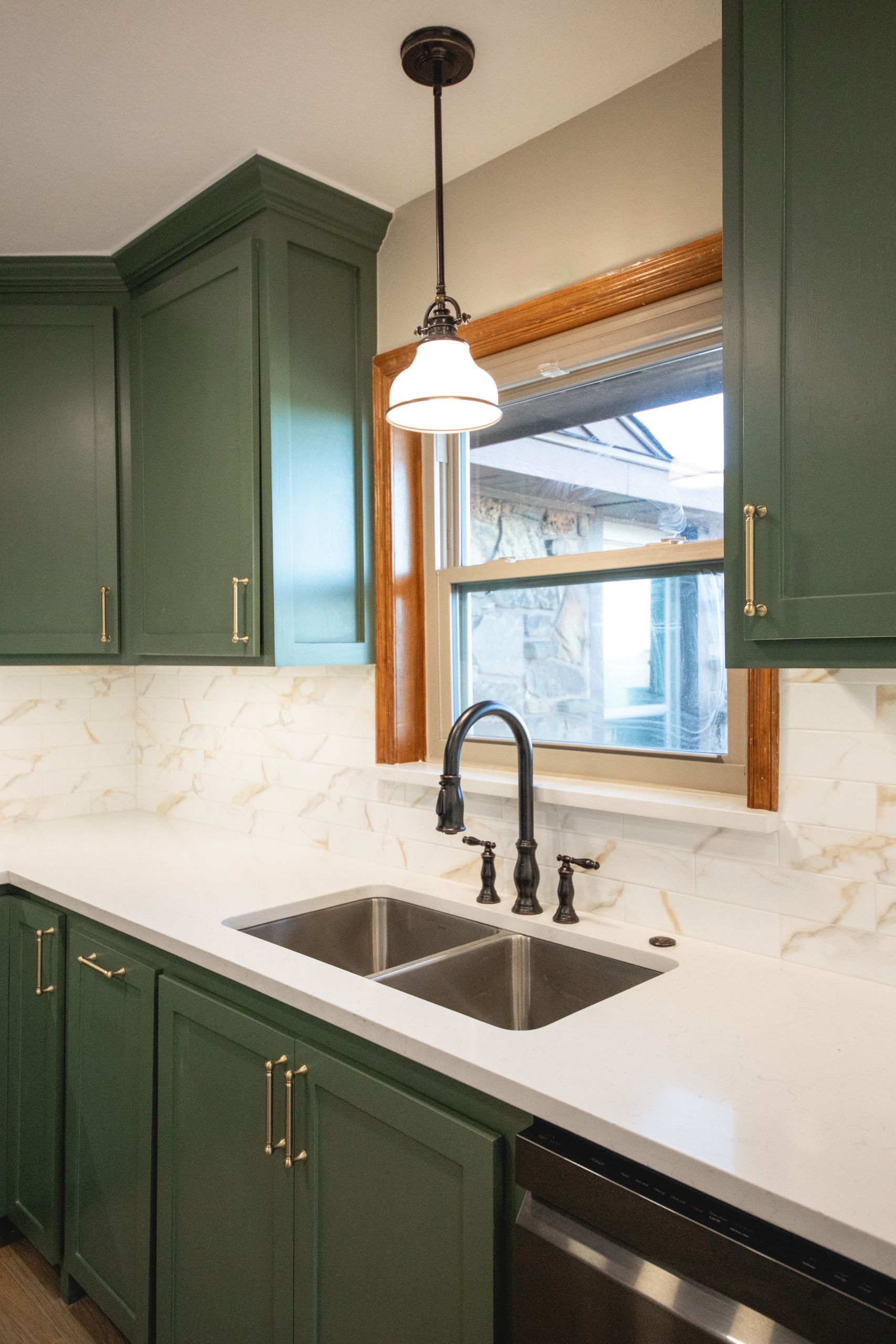Close-up of a double kitchen sink beneath a window with green cabinets and marble backsplash.
