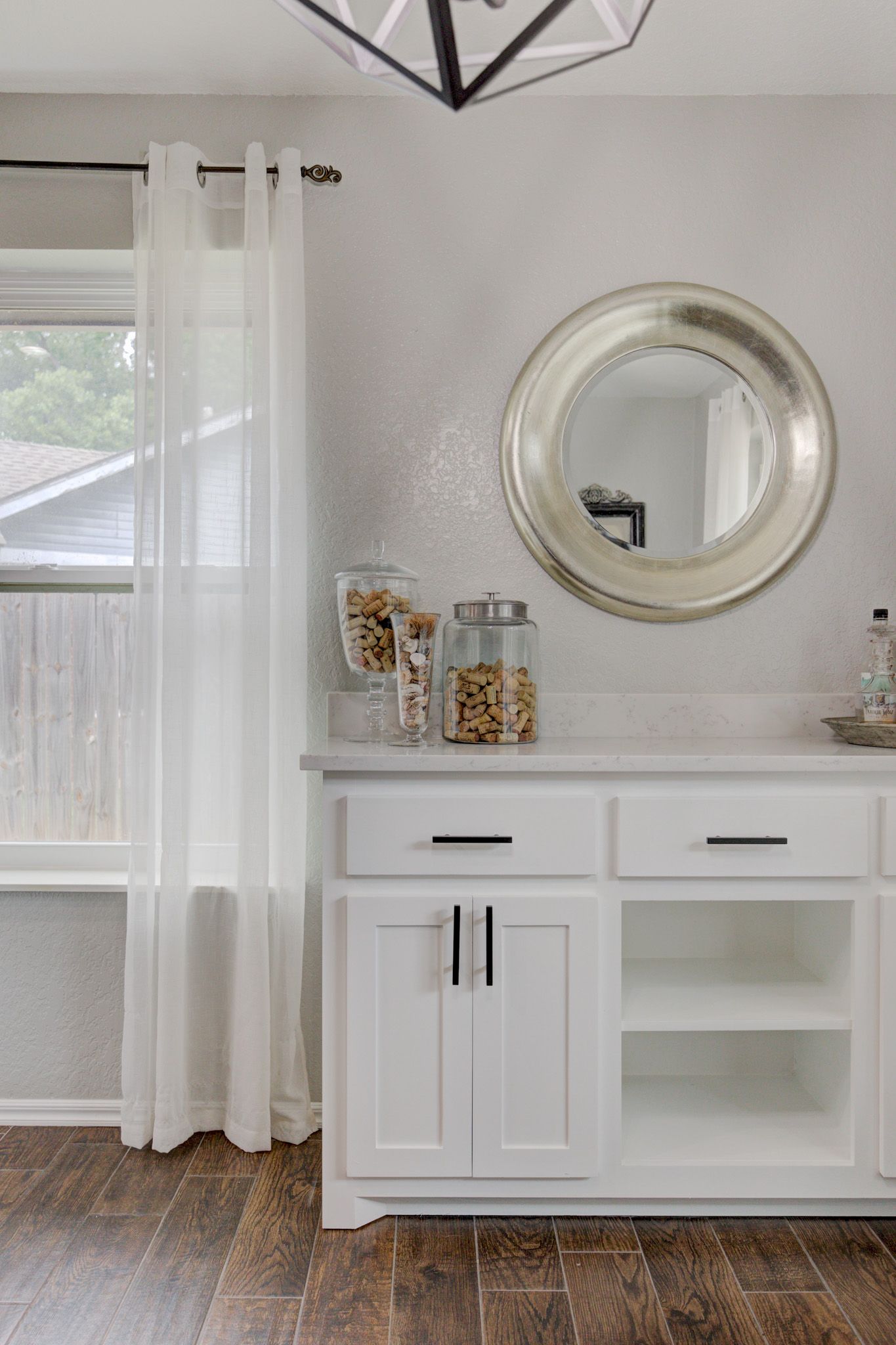 Decorative white cabinet setup with glass jars of corks, a silver round mirror, and sheer curtains.
