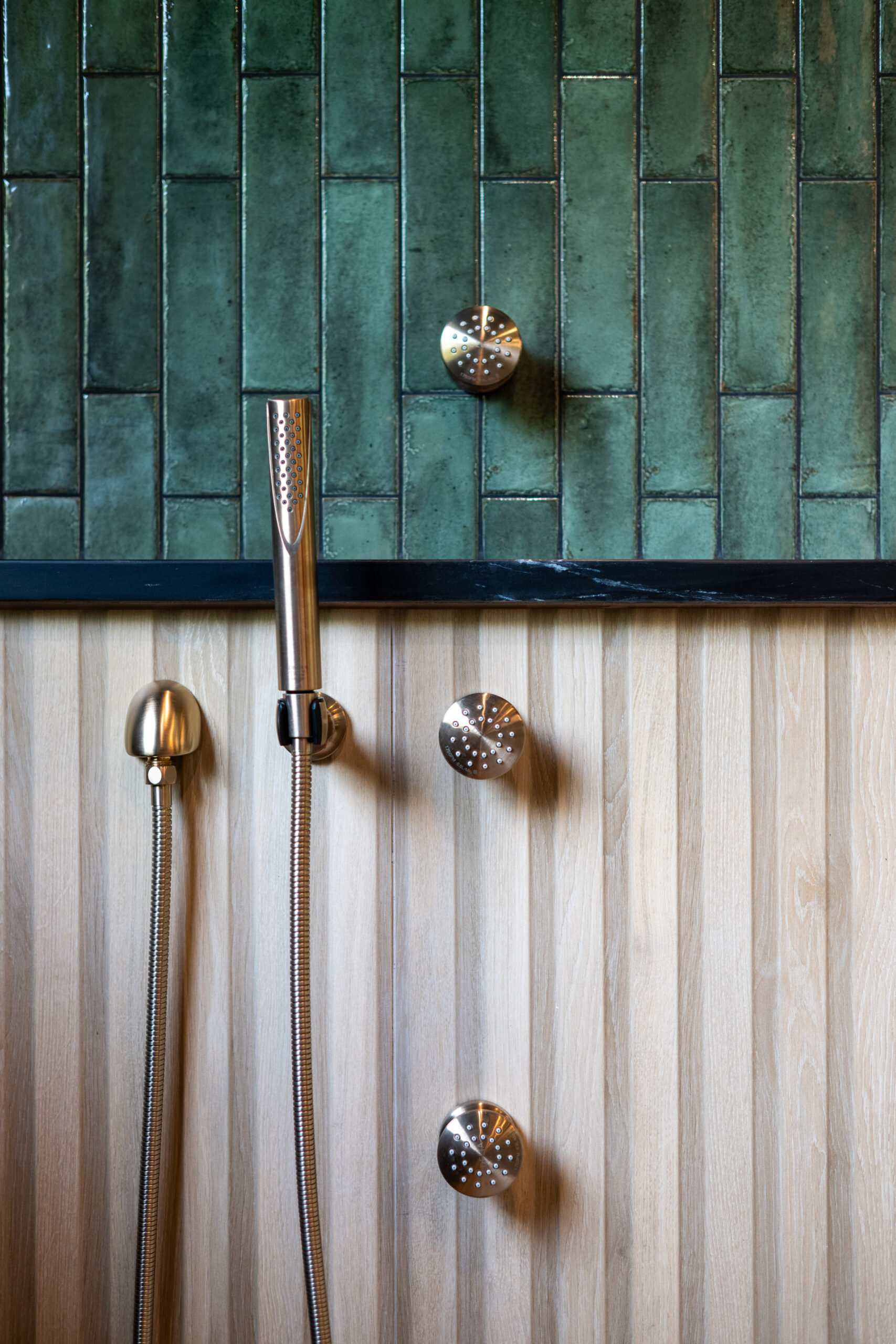 Close-up of bronze shower fixtures against green tile and wood panel walls.