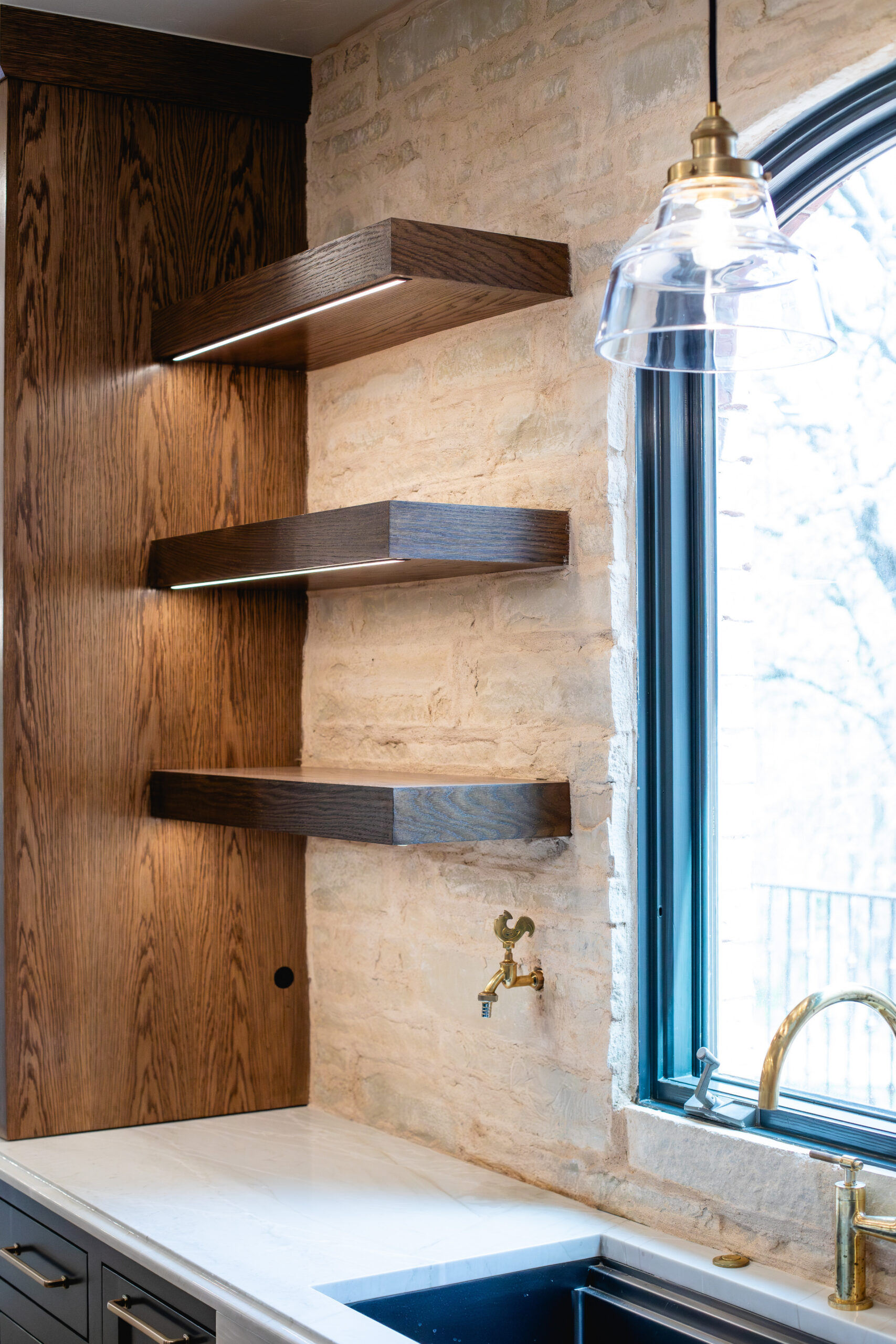 Wood floating shelves with underlighting beside a stone kitchen wall and window.